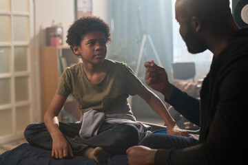Portrait of young Black boy talking to father sitting on bed together in affectionate setting