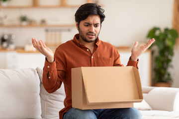 A man with a confused expression opens a cardboard box while sitting on a white couch in a living room. He looks disappointed, as if he did not receive the item he expected.