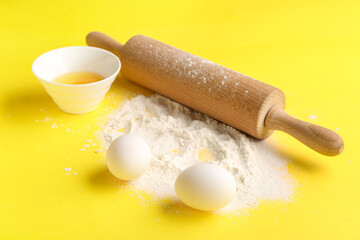 Chicken eggs, flour and rolling pin on yellow background