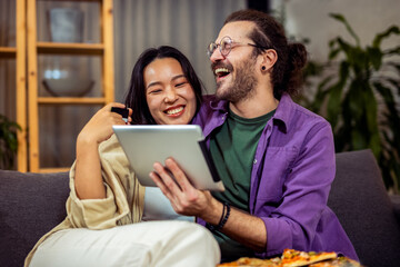 A couple, an Asian woman and a white man, is comfortably seated in their cozy living room. They are engaged in making an online payment together,