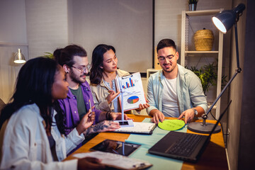 A group of four young people is sitting together at home, collaborating on a project using a laptop, with focused and engaged expressions.