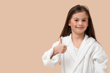 Cute little girl in bathrobe with under-eye patches showing thumb-up gesture on beige background