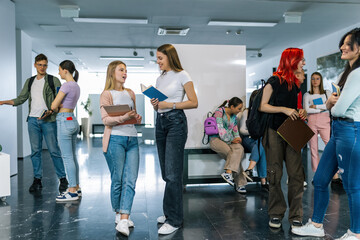 Two best friends, students, walk in the hall of school surrounded with their friends. They talk about the book.