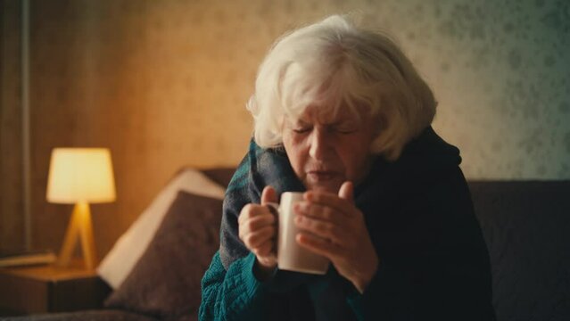 Sad elderly woman wrapped in blanket drinking hot beverage, expensive utilities