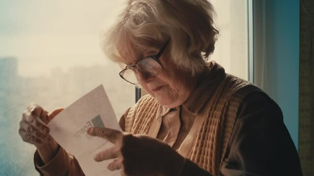 Old woman in glasses reading utility bill letter, shocked by high energy costs