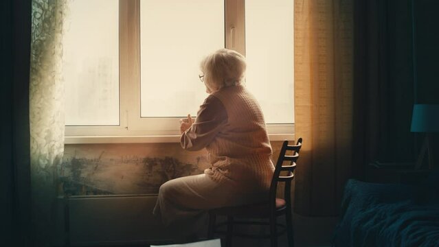 Upset elderly woman looks out the window and drinks tea, loneliness at old age