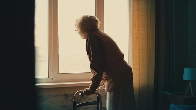 Unhappy senior woman with walking frame looking out the window, feeling lonely