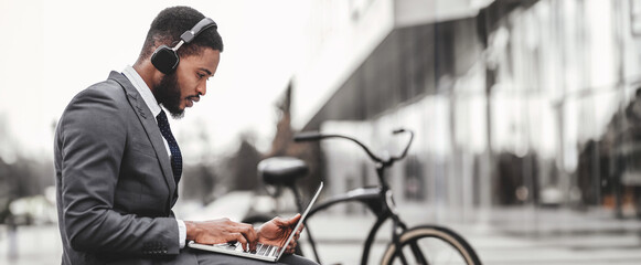 African American businessman using headphones and laptop outdoors, sitting on concrete bench at downtown, bicycle by him, side view, copy space