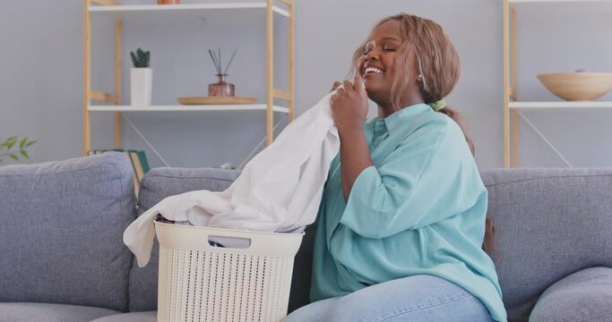 Happy African American woman takes clean, perfectly white shirt out of laundry basket, smells pleasant fabric conditioner, checks for any stains left and remains satisfied. Washing clothes concept