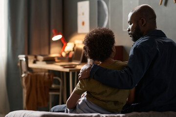 Back view portrait of caring single father embracing son sitting on bed together and talking copy...