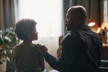 Back view portrait of caring single father comforting young son sitting on bed together against window light