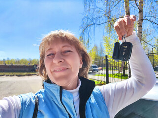 A middle-aged woman takes a selfie with her car keys while standing outdoors on a sunny day