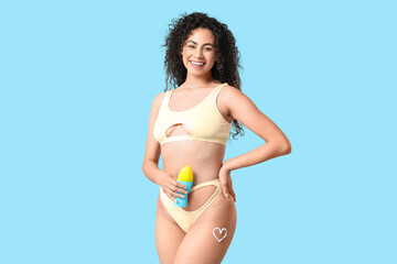 Beautiful young African-American woman in swimsuit with heart made of sunscreen cream on her leg against blue background