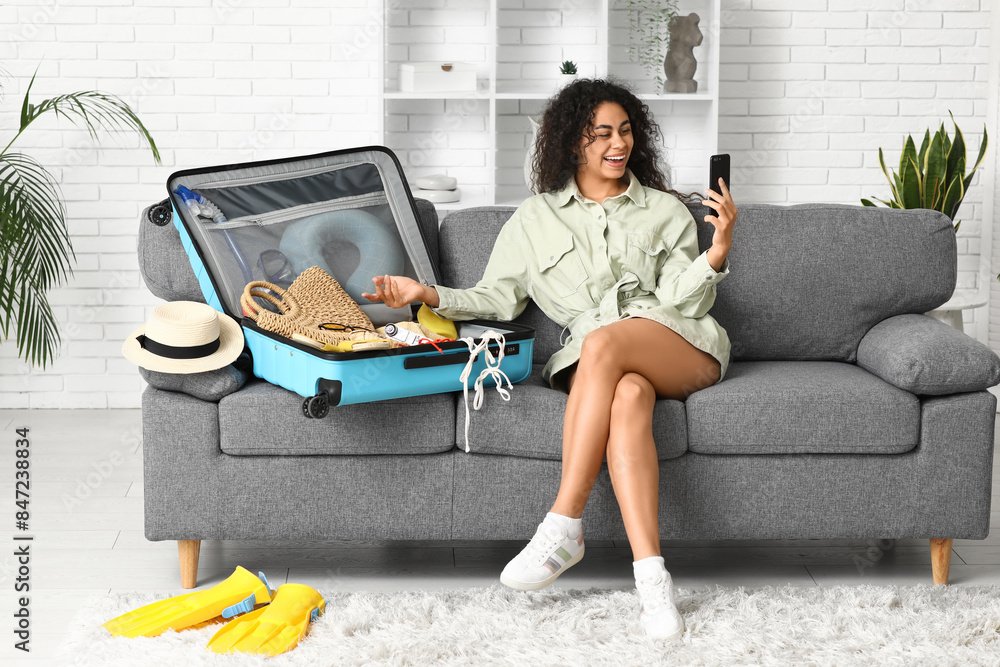 Wall mural beautiful young happy woman with mobile phone unpacking summer clothes from suitcase at home