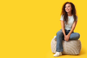 Beautiful young happy African-American woman resting on pouf against yellow background