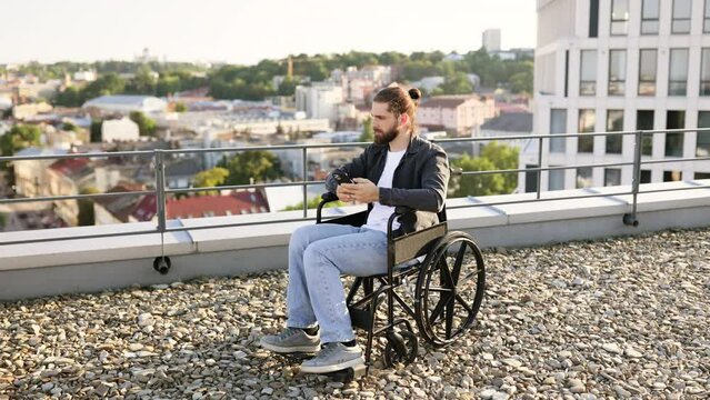 Young Caucasian man in wheelchair using smartphone outdoors on urban rooftop, typing or browsing. Independence, technology, and accessibility in modern city life.
