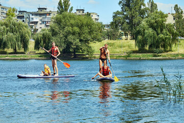 Family enjoying a sunny day paddleboarding on a lake, with lush green trees and apartment buildings in the background.
