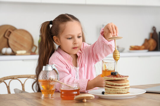 Cute little girl pouring honey on pancakes at table in kitchen
