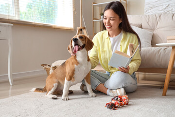 Young woman holding book and playing with cute Beagle dog on floor in living room