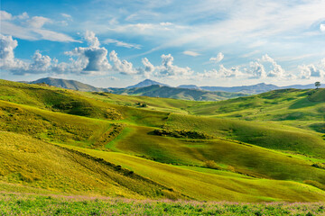 Obraz premium beautiful rural view at summer or spring season fields and hills with rustic grassland and nice mountains with blue cloudy sky on background of countyside landscape