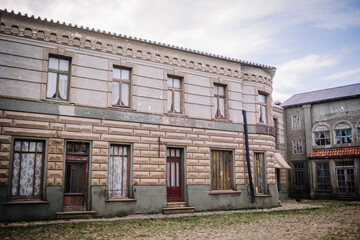 Dobele, Latvia - June 7, 2024 - An old, weathered building with a brick facade, boarded-up windows, and lace curtains, standing on a cobblestone street under a cloudy sky.