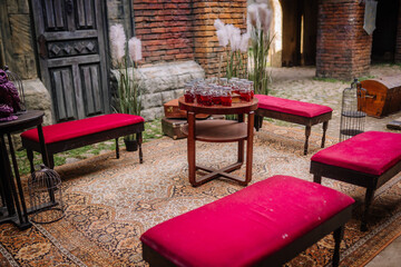 Dobele, Latvia - June 7, 2024 - Outdoor seating area with red cushioned benches, a round table with jars, and potted plants on a decorative rug, set against a brick wall.