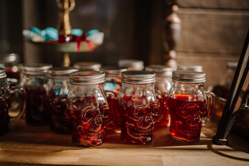 Dobele, Latvia - June 7, 2024 - Skull-shaped glass jars filled with a red beverage on a wooden surface, with blurred blue and red sweets in the background.