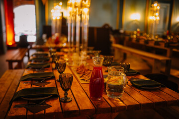 Dobele, Latvia - June 7, 2024 - Elegant wooden dining table set with glassware, cutlery, and pitchers of drinks, under warm candlelight in a cozy setting. Copy space.