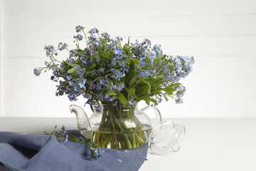 Bouquet of beautiful forget-me-not flowers in glass teapot and blue cloth on white table