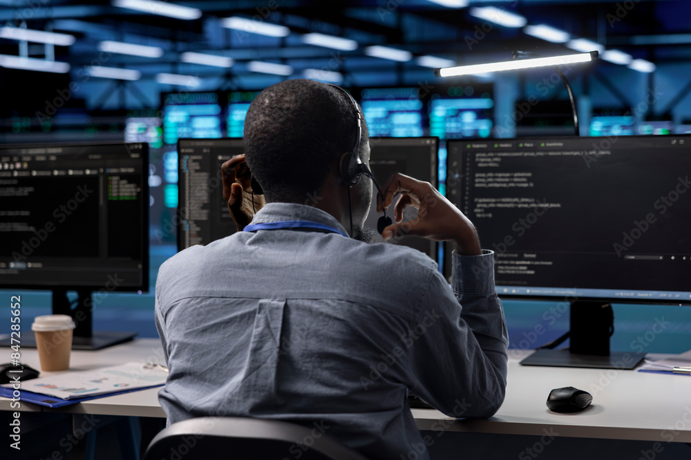Poster technician in server room managing call center tasks, speaking into headset while monitoring multipl
