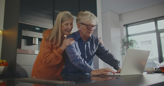 Camera moving around older Caucasian couple. People standing in middle of kitchen. Wife hugging husband from behind. Man operating laptop. Writing sentences or email to relative. Remote communication.