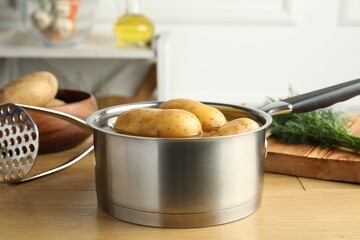 Raw potatoes in saucepan with water, pusher and dill on wooden table