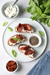 Delicious ricotta bruschettas with sun dried tomatoes, basil and milled pepper on light table, flat lay