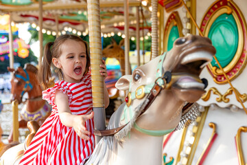 Adorable little brunette girl in summer red-white dress at amusement park having a ride on the merry-go-round. Child girl has fun outdoor on sunny summer day. Entertainment concept