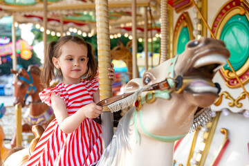 Adorable little brunette girl in summer red-white dress at amusement park having a ride on the...