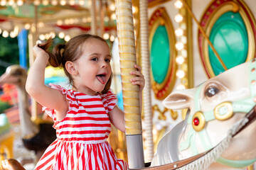Adorable little brunette girl in summer red-white dress at amusement park having a ride on the...