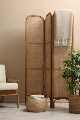 Folding screen, blanket, armchair and green houseplant near beige wall indoors