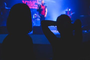 A crowded concert hall arena with scene stage lights with musicians band on a stage at the venue, rock show performance, with concert-goers attendees, audience on dance floor during concert festival