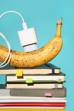 Phone charger plugged into a banana on top of colorful notebooks. 