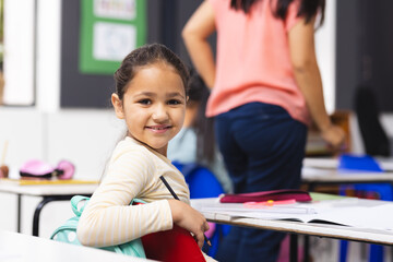 Biracial girl sitting at desk in classroom, smiling at camera