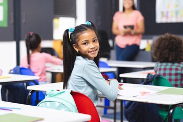 Biracial girl sitting at desk in classroom, smiling at camera