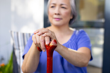 Asian senior woman holding cane with both hands, looking thoughtful, at home