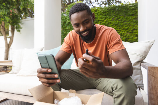 Young African American man smiling at his smartphone, holding a small item, at home