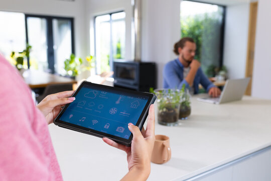 Young biracial woman using tablet with smart home app, Caucasian man working on laptop - Powered by Adobe