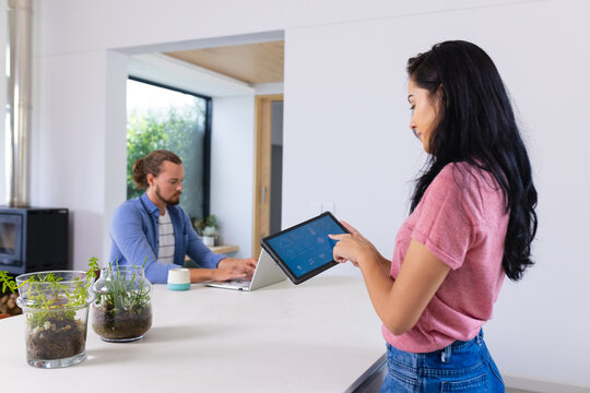 Young biracial woman using tablet with smart home, caucasian man working on laptop