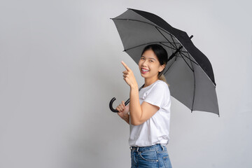 Happy Asian woman standing holding a umbrella and pointing at copy space on gray background.