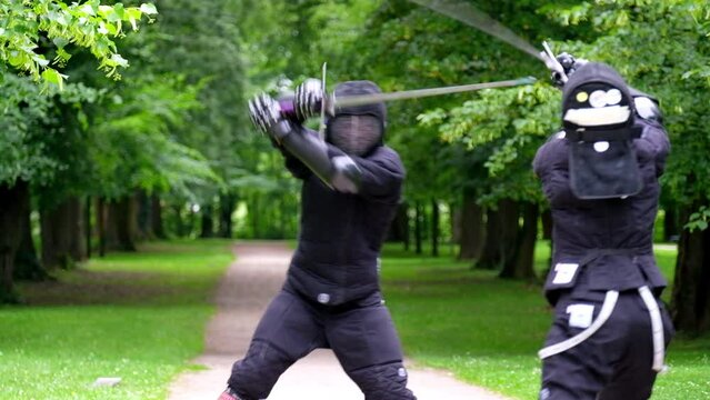 Two fencers dressed in black protective uniforms, helmets with face masks are fencing in city park using long medieval metal historical swords, practicing before competition 4K video.