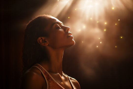 Woman of faith with eyes closed in prayer is blessed with divine light shining on her face. She looks full of devotion & grace, experiencing the bliss of a mystical experience, a spiritual awakening.