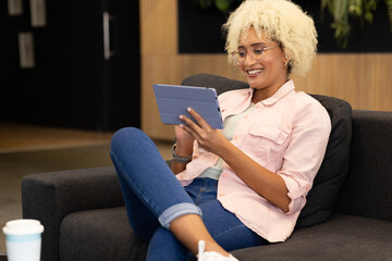 Young biracial woman, team member, using tablet and smiling in business office lounge area
