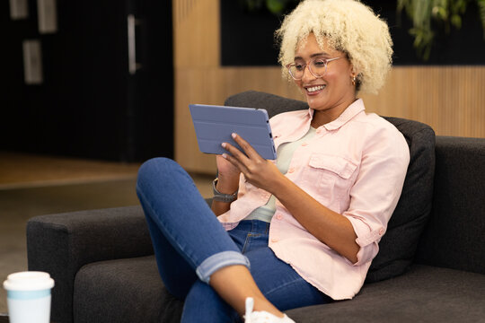 Young biracial woman, team member, using tablet and smiling in business office lounge area